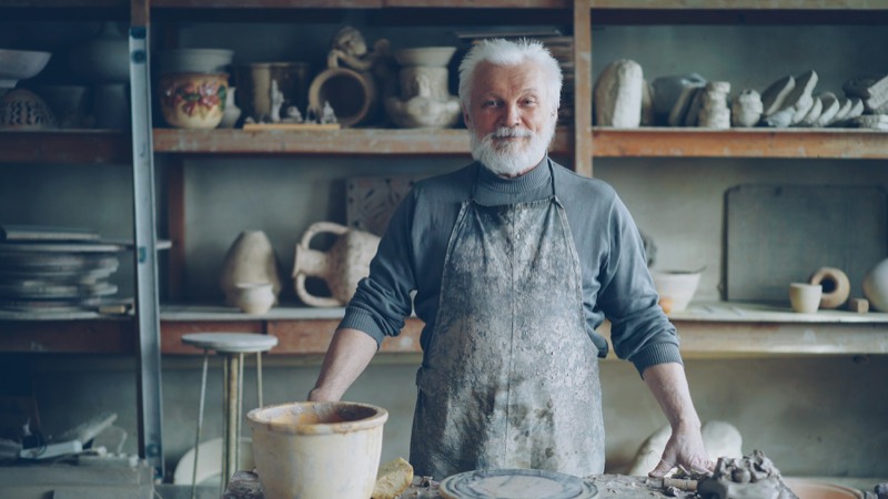 Pottenbakker met grijze baard en bestoven schort aan zijn werktafel, achter hem rekken vol aardewerk
