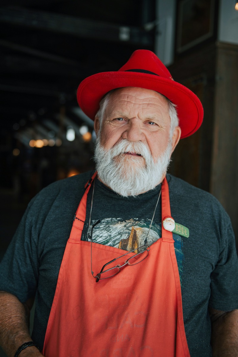 Oudere man met witte baard, rode hoed en oranje schort in zijn werkplaats